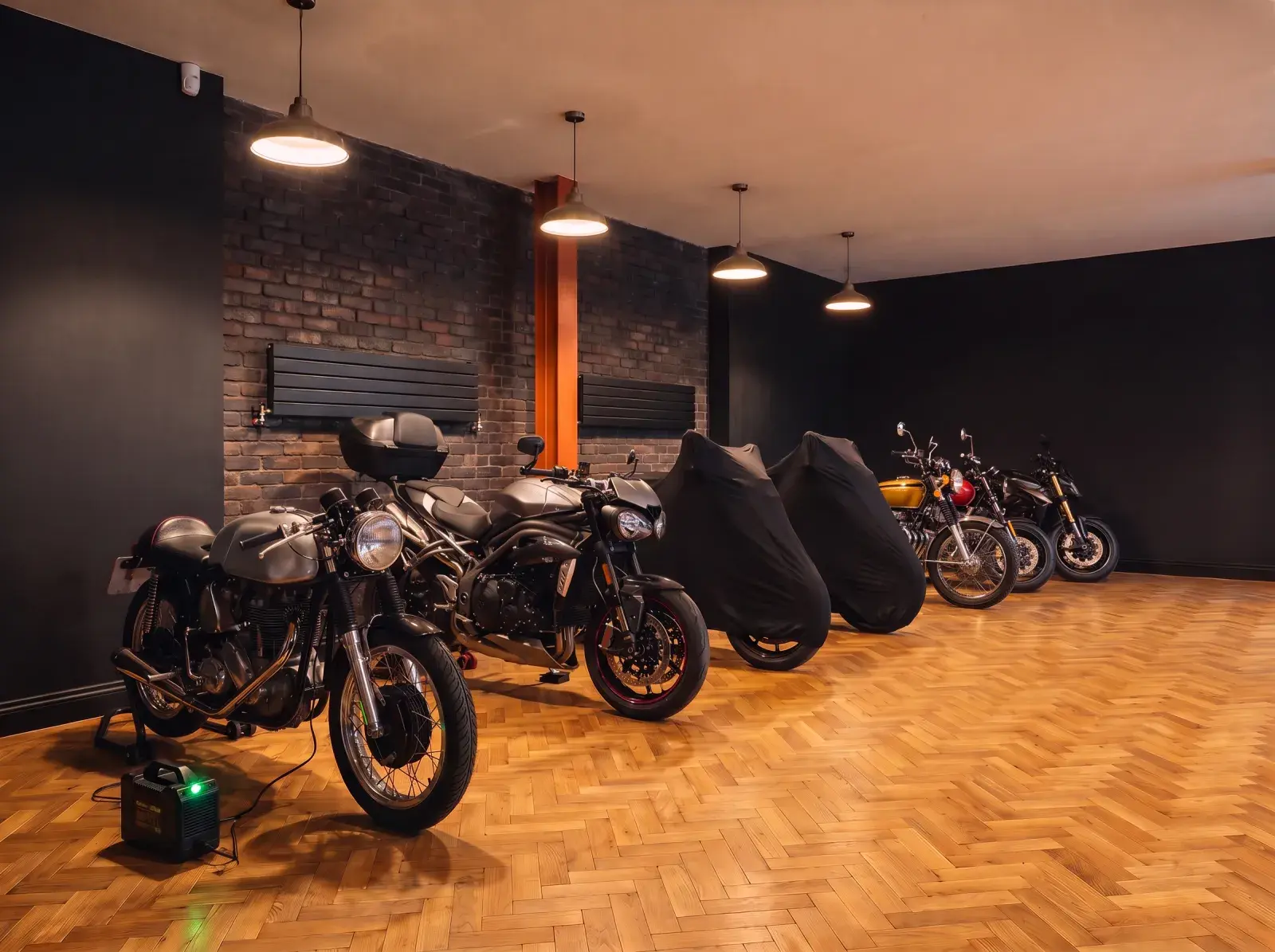 Motorräder in einer Reihe auf Holzoptik-Boden im Fischgrätmuster in einer warm beleuchteten Halle, zwei unter Abdeckhauben, am vorderen hängt ein Batterie-Ladegerät